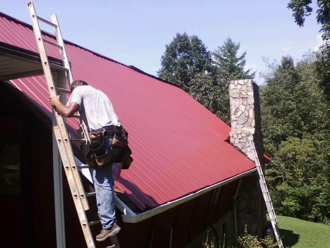 Roofer on a ladder beside a red metal roof and stone chimney — Soffit & Fascia Repair by Roofing Brothers in Forest Hills
